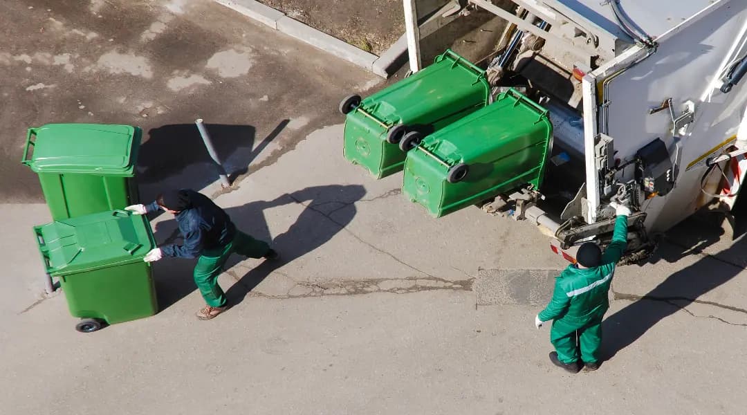 two people working for waste trucks putting waste bins on the back of waste trucks.