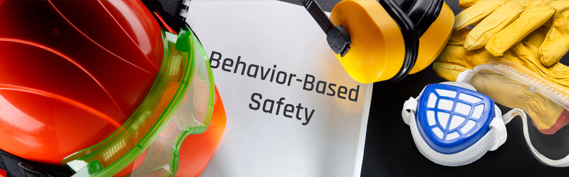 various safety gear items, including a red hard hat, yellow ear protectors, yellow safety gloves, and a white respirator mask with blue accents, arranged around a document titled "Behavior-Based Safety." The safety equipment is placed on a dark surface, highlighting the importance of safety measures in the workplace.